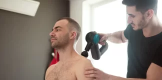 Fizjoterapeuta wykonuje zabieg urządzeniem na barku siedzącego mężczyzny.